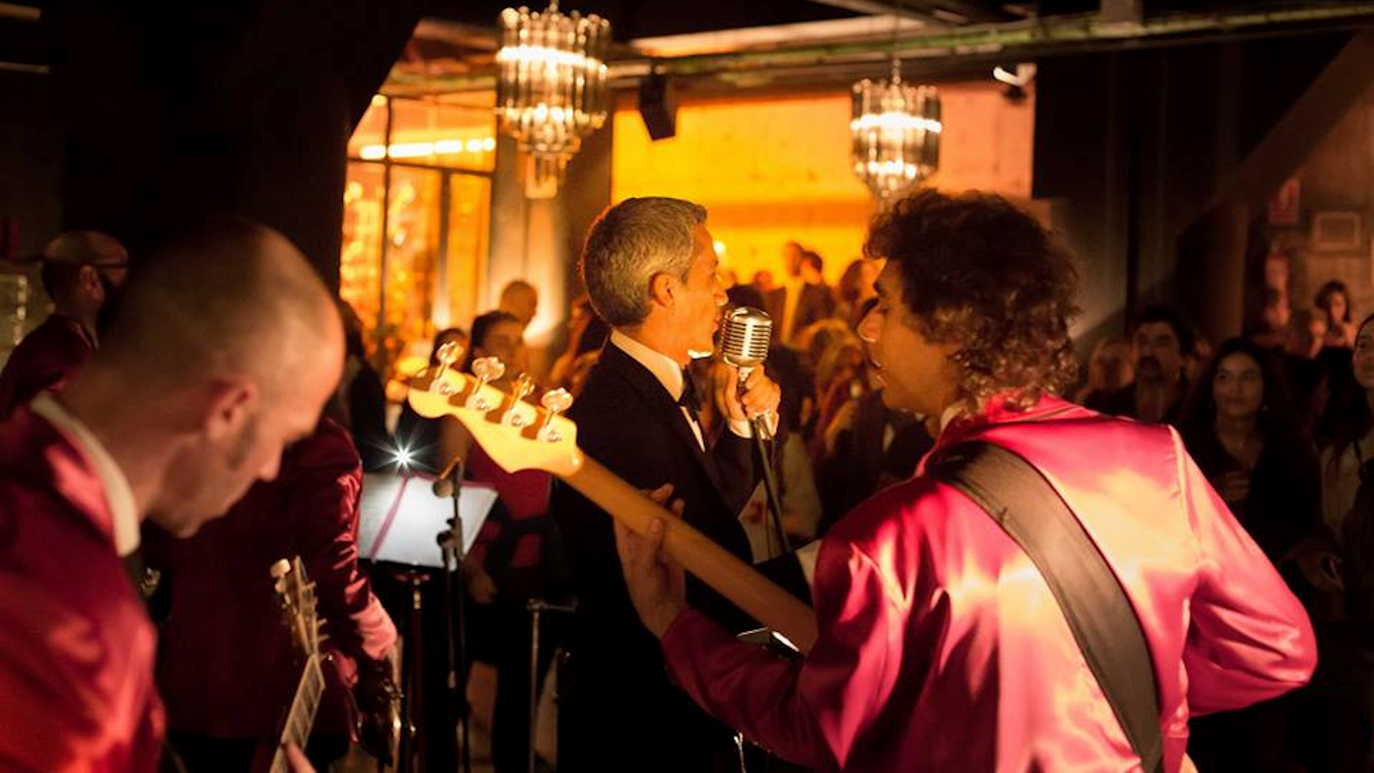 veduta di spalle della band che suona durante un concerto. Davanti a loro il pubblico in un locale elegante, con lampadari di cristallo.